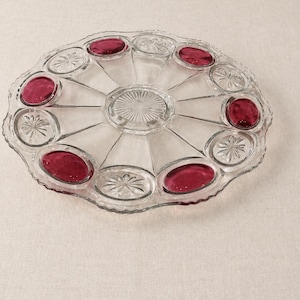 May include: A round clear glass serving platter featuring a scalloped edge and a central sunburst pattern. The platter is divided into twelve alternating sections, with six smooth deep magenta oval inserts and six clear glass starburst panels.