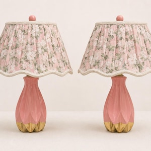 May include: A pair of table lamps featuring pink, fluted ceramic bases with gold-toned metallic accents at the bottom. Each lamp is topped with a pleated, scalloped-edge lampshade decorated with a pink and green floral pattern on a light background.