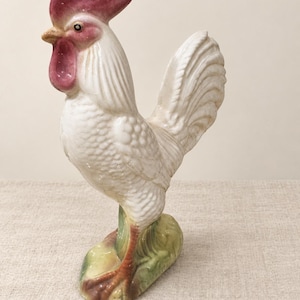 May include: A ceramic rooster figurine featuring a white body with textured feather details, a prominent red comb and wattle, and a yellow beak. The bird stands on a green and brown base, with its head turned slightly to the left, showcasing a glossy finish.