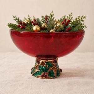 May include: A deep red glass bowl with a textured surface, supported by a decorative metal pedestal base featuring green holly leaves, gold outlines, and small red berries, filled with evergreen sprigs and gold ornaments.
