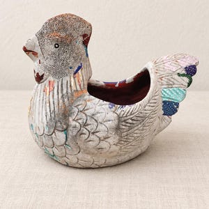 May include: A ceramic chicken-shaped planter with a weathered white finish and textured feather details. The piece features accents of orange, blue, and green paint on the wings and tail, with a deep red interior and a hollow center for planting.