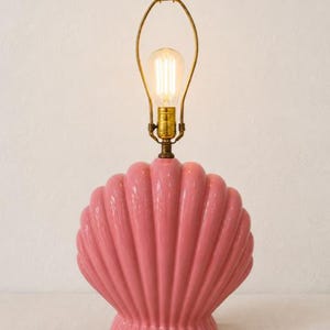 May include: A glossy pink ceramic table lamp shaped like a large scallop shell. The lamp features a brass-colored metal harp and socket assembly extending from the top, holding a single clear glass Edison-style light bulb with visible filaments.