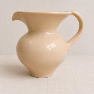 May include: A solid light beige ceramic pitcher with a smooth, glossy finish. The vessel features a rounded, bulbous body, a wide flared rim with a distinct pouring spout, and a curved handle attached to the side for easy gripping and pouring.