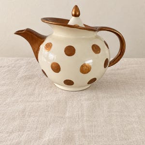 May include: A ceramic teapot with a cream-colored body featuring metallic bronze polka dots. The teapot includes a matching lid with a bronze finial, a bronze-colored spout, and a curved bronze handle. The finish on the bronze elements is glossy.