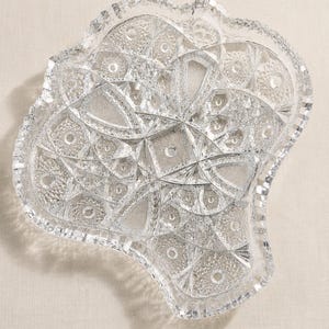 May include: A clear, colorless glass serving dish with an irregular, organic shape. The surface features intricate, geometric cut patterns including cross-hatching, starburst motifs, and faceted edges that create a highly reflective, textured appearance.