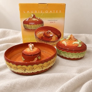 May include: A Laurie Gates 4-piece serve set Santa Fe featuring a large round ceramic bowl with a central raised section and a matching lidded dish. The pieces are painted in shades of orange, red, and yellow with green chili pepper accents.