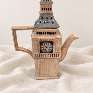 May include: A ceramic teapot shaped like the Elizabeth Tower, featuring a beige body with a square clock face, a handle, and a spout. The top is a removable lid with a blue-grey roof, gold accents, and small cut-out details on the tower structure.