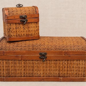 May include: Two rectangular storage chests with arched lids, constructed from woven brown material with smooth wooden frames. Each chest features a dark metal latch on the front and a small metal handle on the top of the smaller chest.