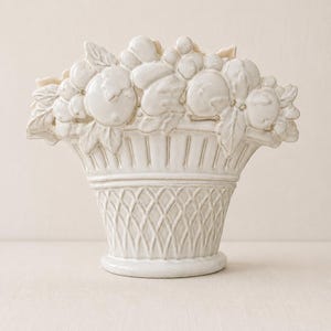 May include: A cream-colored ceramic basket vase featuring a relief design of assorted fruits and leaves along the top rim. The body of the vessel displays a textured lattice pattern on the lower section and vertical fluting above the base.