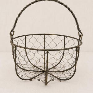 May include: A round wire basket with a dark bronze finish, featuring a hexagonal mesh pattern and a sturdy frame. The basket includes a tall, arched handle with a coiled metal design, attached to the sides with small loops for easy carrying.