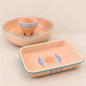 May include: A peach-colored ceramic set consisting of a large round bowl with a scalloped rim, a small three-legged bowl inside it, and a rectangular baking dish. Each piece features a teal rim and geometric patterns in blue and orange.