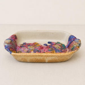 May include: A rectangular ceramic baking dish with rounded corners and small side handles. The exterior has a speckled tan finish, while the interior features a vibrant, abstract glaze pattern in shades of magenta, deep blue, and golden yellow.