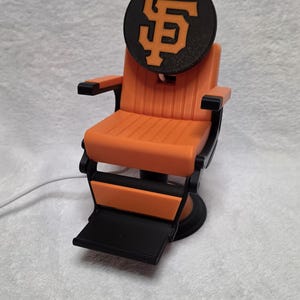 Może przedstawiać: Miniaturowy fotel fryzjerski w kolorze pomarańczowo-czarnym z okrągłym zagłówkiem z pomarańczowym logo San Francisco Giants. Fotel posiada żebrowane siedzisko, czarne podłokietniki, podnóżek oraz okrągłą podstawę z wystającym białym przewodem zasilającym.