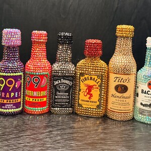 Puede incluir: Una colección de once botellas de licor en miniatura, cada una totalmente incrustada con pequeños diamantes de imitación brillantes en colores que combinan con sus etiquetas. Incluye marcas como 99, Jack Daniel's, Fireball, Tito's, Bacardi, Smirnoff y Don Julio.