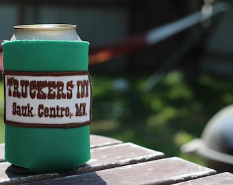 May include: A green can cooler with a brown patch that says "Truckers Inn Sauk Centre, MN."