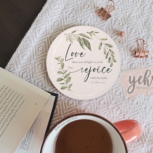 Può includere: Un sottobicchiere rotondo, rosa e bianco con un design a foglie verdi. Il sottobicchiere presenta il testo "L'amore non si compiace del male, ma si rallegra della verità. 1 Corinzi 13:6".