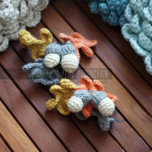 May include: Four crocheted amigurumi toys in the shape of fish. The fish are in shades of grey, orange, and yellow. The fish are sitting on a wooden surface.