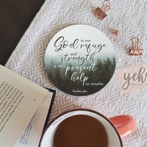 May include: A round, white coaster with a forest scene and the text "God is our refuge and strength, a very present help in trouble. Psalm 46:1". The coaster has a green and white watercolor background with a light blue fog effect. The text is in black cursive font.