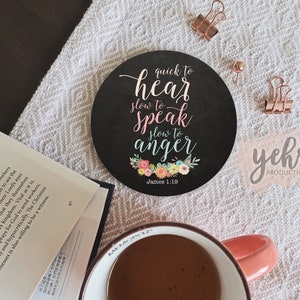 Può includere: Un sottobicchiere rotondo nero con il testo "quick to hear, slow to speak, slow to anger" in un carattere corsivo, con accenti floreali. Una tazza di caffè, un libro e articoli per ufficio sono anche nell'immagine.