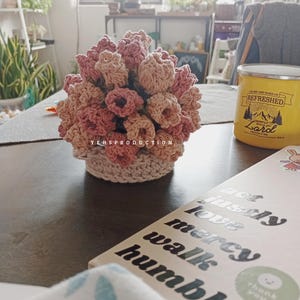 May include: A handmade crochet floral arrangement in shades of pink and beige. The flowers are clustered together in a round shape, sitting in a beige crochet basket. The text "YEHSPRODUCTION" is visible. A yellow mug is in the background.