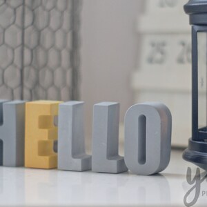 Puede incluir: La palabra "HELLO" escrita en letras de bloque grises y amarillas. Las letras están sobre una superficie blanca.