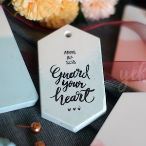 Pode incluir: Um conjunto de três etiquetas de presente de cerâmica branca com um design degradê em azul claro, rosa e branco. A etiqueta do centro tem o texto "Above all else Guard your heart" escrito em caligrafia preta.