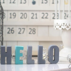May include: Grey and teal painted wooden letters spelling "HELLO" on a white surface with fairy lights.