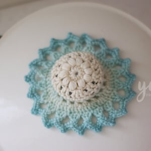 Puede incluir: Un posavasos de ganchillo blanco con un diseño degradado azul y blanco. El posavasos está sobre una superficie blanca.