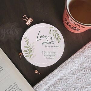 May include: A round pink coaster with a white border and green leaves. The coaster features the text "Love is patient. Love is kind. It does not envy, it does not boast, it is not proud. (1 Corinthians 13:4)"