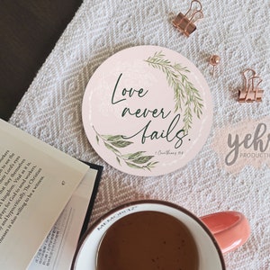 Può includere: Un sottobicchiere rotondo rosa e verde con il testo "L'amore non fallisce mai." e un design floreale. Il testo "1 Corinzi 13:8" è incluso anche sul sottobicchiere.