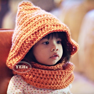Puede incluir: Un conjunto de capucha y cuello de ganchillo naranja. La capucha tiene una parte superior puntiaguda y enmarca la cara. El cuello es de color naranja a juego. El conjunto está hecho de un hilo grueso y es probablemente para un niño. La imagen incluye el texto "YEHs".