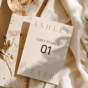 以下が含まれることがあります： セージグリーンのセリフ体でASHLEYとPETERの名前、ダークグリーンの文字でTABLE NUMBER 01、そしてNORTH VANCOUVER, BC SEPTEMBER 23, 2026と記された、クリーム色の長方形のウェディングテーブルナンバーカード。