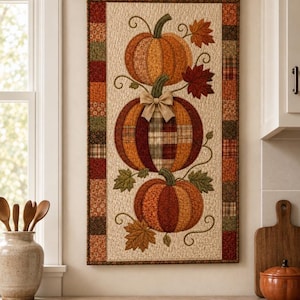 May include: A rectangular fabric wall hanging featuring a vertical stack of three quilted pumpkins in shades of orange, brown, and plaid. The center pumpkin is adorned with a tan fabric bow, and the design includes embroidered leaves and vines.