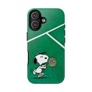 Op de afbeelding: Een groene smartphonehoes met een witte tennisbaanlijn-afbeelding en een cartoonillustratie van een witte hond met zwarte oren die een blauwe hoofdband draagt en een groen tennisracket vasthoudt met een gele bal in het midden van het racket.
