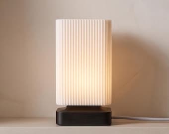 May include: A rectangular table lamp featuring a white, vertically ribbed glass shade that emits a soft glow. The shade is supported by a solid, dark brown wooden base with a smooth, matte finish. A light-colored power cord extends from the side.