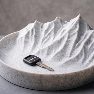 May include: A circular white tray with a speckled gray finish featuring a three-dimensional mountain range relief on one side. A black Mazda car key rests on the flat surface of the tray, which is designed to hold small items like keys.