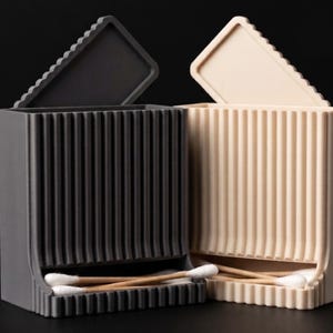 May include: Two rectangular storage containers with vertical ribbed textures, one dark gray and one beige. Each container features an open-top design with a hinged lid and a bottom dispensing slot, currently holding several cotton swabs with white tips.