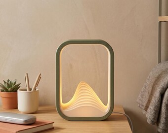 May include: A modern, rectangular table lamp with a sage green frame and a layered, wave-like design that emits a warm glow. The lamp is on a wooden surface, next to a small potted plant, pens, and a book.