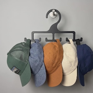 Könnte beinhalten: Ein schwarzer Hut-Organizer-Kleiderbügel aus Kunststoff mit einem großen Haken und fünf einzelnen Halterungen, an denen fünf Baseballkappen in Grün, Blau, Ocker, Creme und Marineblau hängen. Die grüne Kappe hat ein Netzteil und einen Aufnäher.