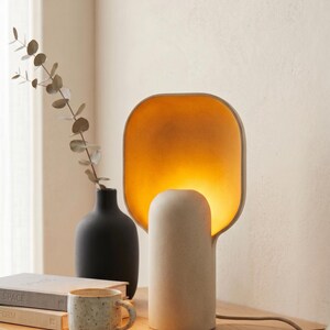 May include: A table lamp with a cylindrical base and a wide, rounded rectangular shade that emits a warm, golden-yellow glow from within. The lamp has a matte, off-white textured finish and is connected to a power cord extending to the right.