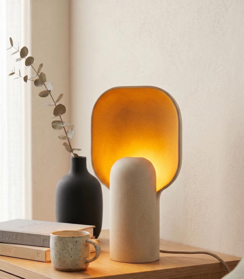 May include: A table lamp with a cylindrical base and a wide, rounded rectangular shade that emits a warm, golden-yellow glow from within. The lamp has a matte, off-white textured finish and is connected to a power cord extending to the right.