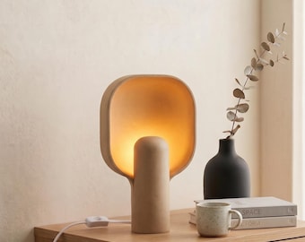 May include: A beige table lamp with a rounded, square-shaped shade and a cylindrical base that emits a warm, soft glow. The lamp features a white power cord with an inline switch, providing a focused light source for a bedside or desk area.
