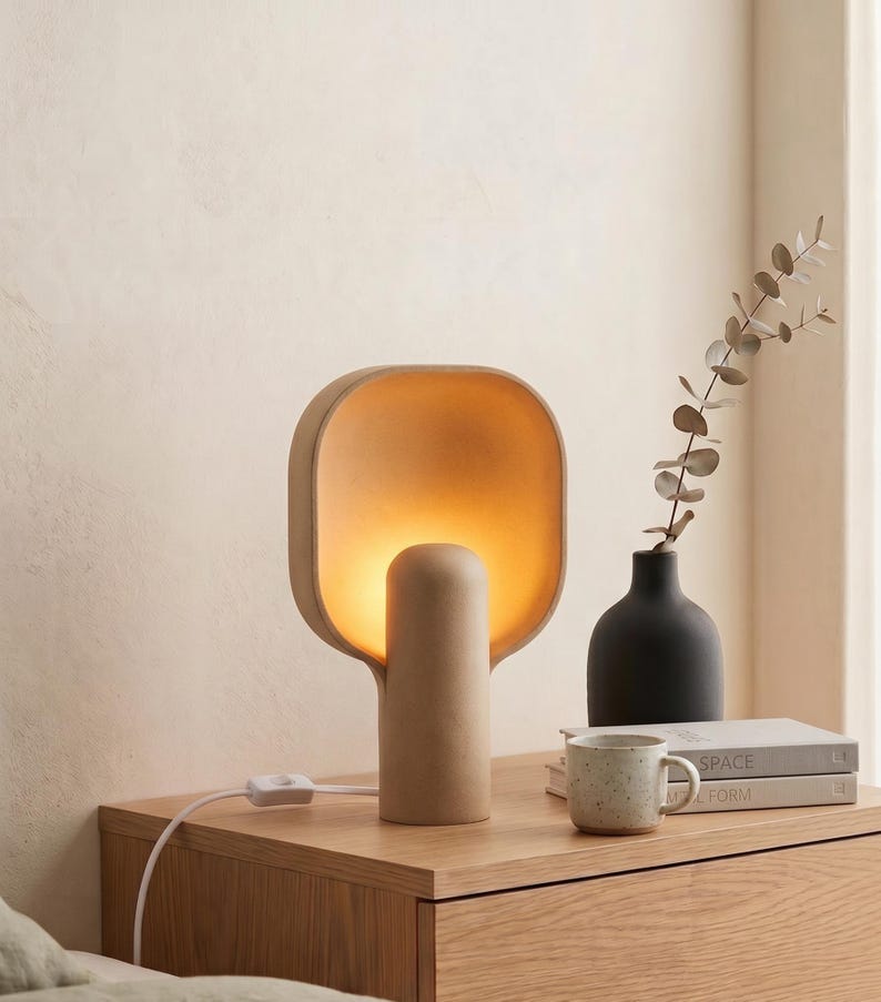 May include: A beige table lamp with a rounded, square-shaped shade and a cylindrical base that emits a warm, soft glow. The lamp features a white power cord with an inline switch, providing a focused light source for a bedside or desk area.