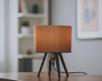 May include: A table lamp featuring a cylindrical, vertically ribbed lampshade in a warm amber tone, supported by three dark, angled wooden legs. The lamp includes a central metallic socket and a black power cord extending from the base.