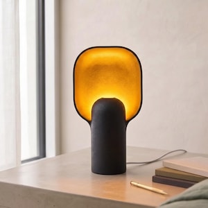 May include: A table lamp with a matte black cylindrical base and a wide, rounded rectangular shade. The interior of the shade features a textured gold finish that glows with a warm, bright light, contrasting against the dark exterior surface.