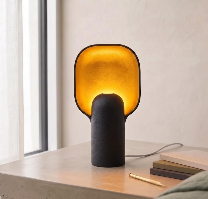 May include: A table lamp with a matte black cylindrical base and a wide, rounded rectangular shade. The interior of the shade features a textured gold finish that glows with a warm, bright light, contrasting against the dark exterior surface.