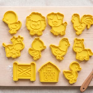 Pode incluir: Um conjunto de dez cortadores de biscoitos de plástico amarelo com várias formas de temática agrícola, incluindo uma ovelha, porco, galo, vaca, cavalo, celeiro, cerca e três aves diferentes, dispostos numa tábua de madeira clara.
