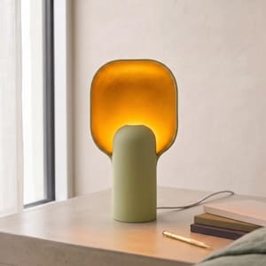 May include: A table lamp with a cylindrical sage green base and a wide, rounded rectangular shade. The interior of the shade features a textured gold finish that glows with warm light, while the exterior matches the matte sage green base.