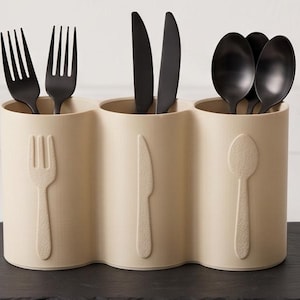 May include: A beige triple-cylinder utensil holder with embossed fork, knife, and spoon icons on the front. The holder contains matte black forks, knives, and spoons organized by type in each section, providing a structured storage solution.