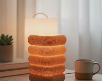 May include: A modern orange table lamp with a stacked, cylindrical design. The lamp has a textured surface and emits a warm glow. It sits on a wooden table, next to a mug, a notebook, and a pen.